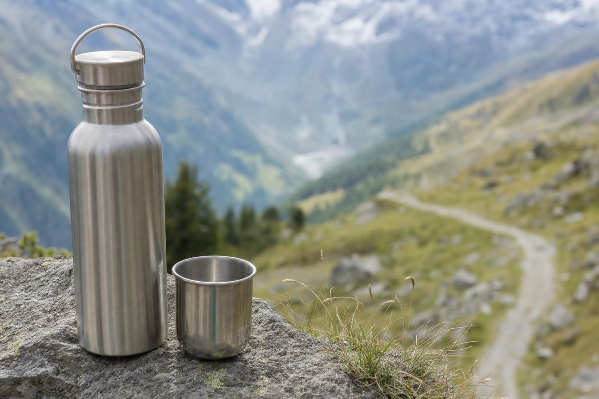 Trinkflasche und Becher auf einem Fels an einem Bergweg, im Hintergrund eine Bergkette
