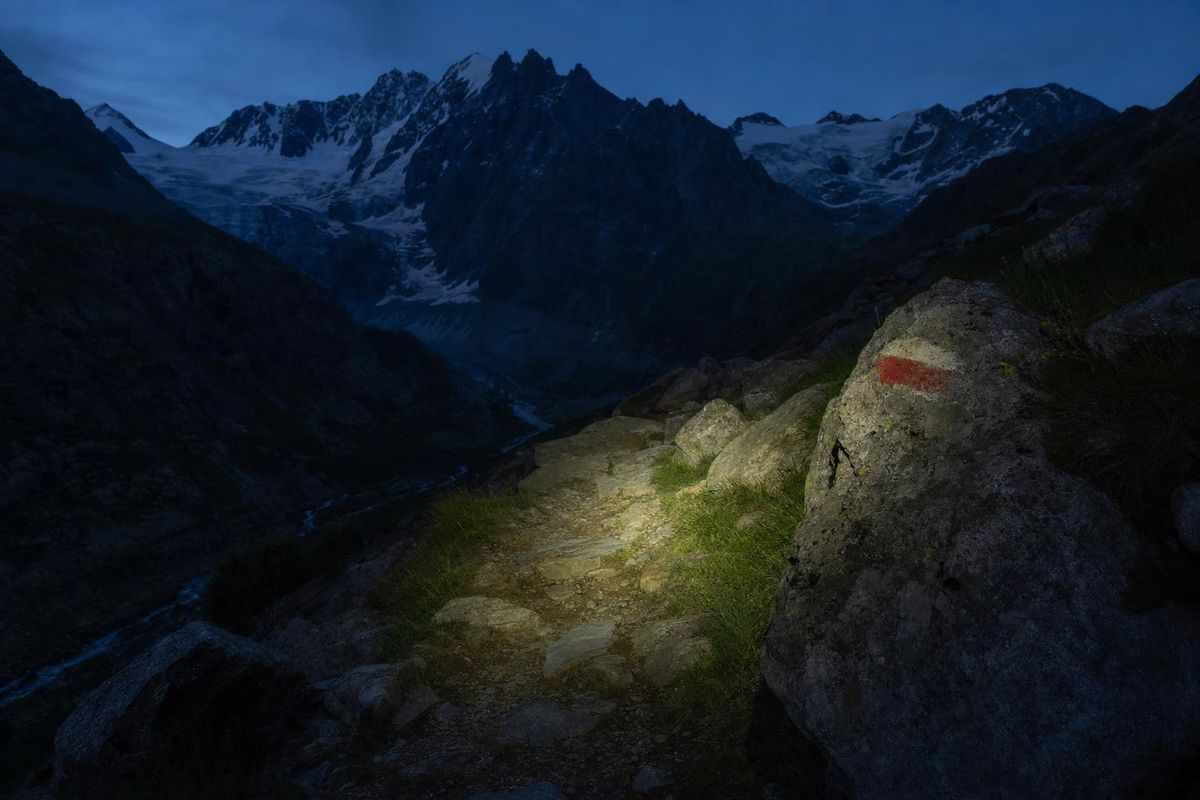 Stirnlampe leuchtet einen schmalen Bergpfad in der Dämmerung aus, roter Markierungspunkt auf einem Fels im Lichtkegel
