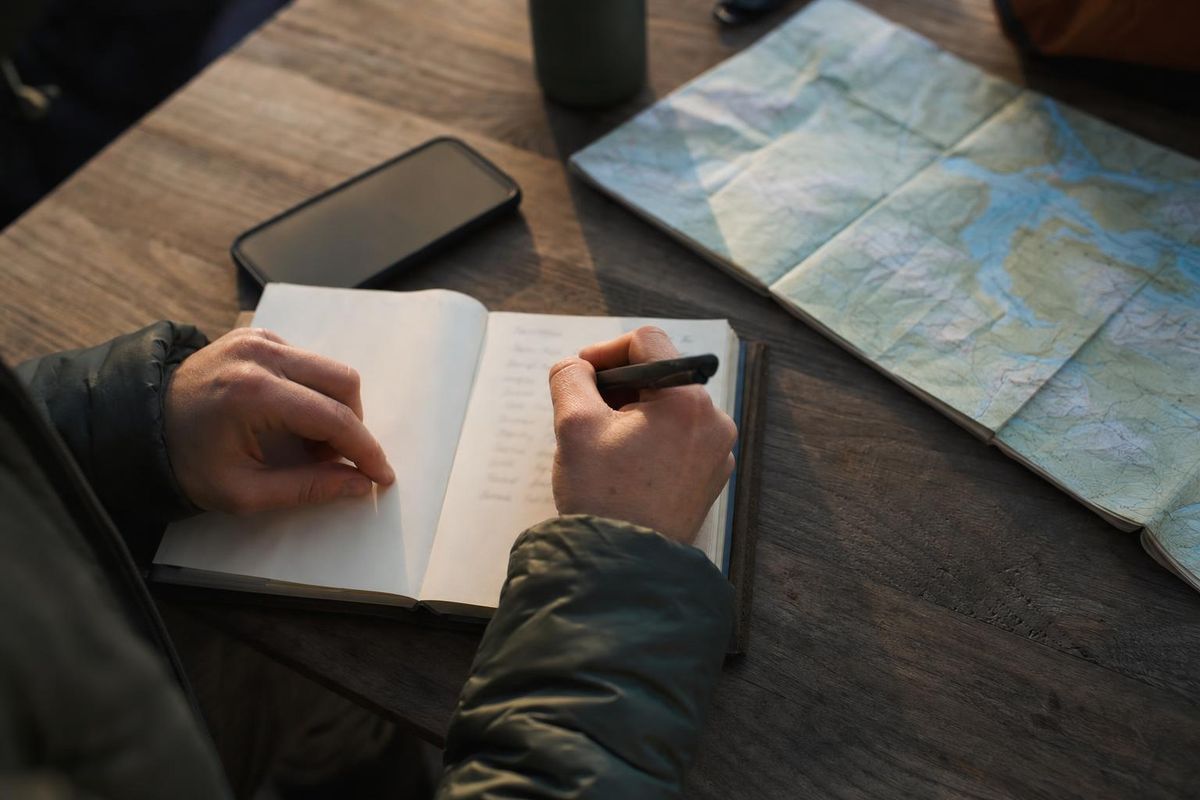 Hand schreibt Namen in ein Notizbuch neben Handy und Wanderkarte