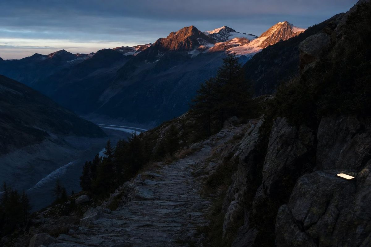Bergweg führt in der Dämmerung ins Tal, letzte Sonne auf den Gipfeln