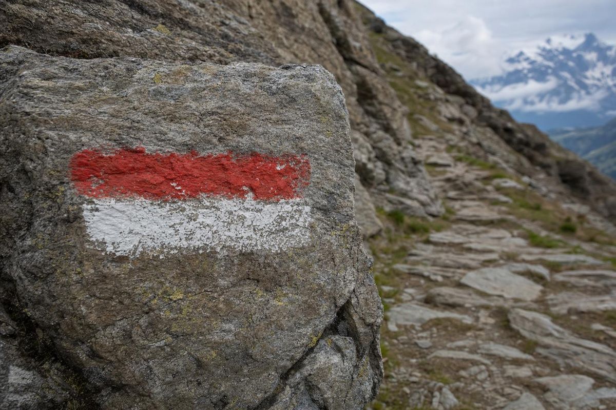 Wegweiser mit rot-weißer Markierung an einem Felsen, dahinter geht ein Steig steil bergauf