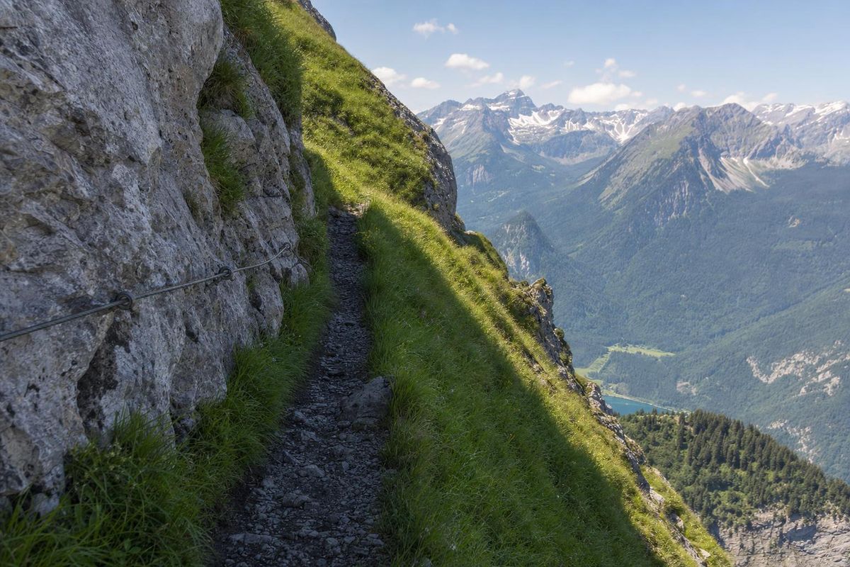 Schmaler Bergpfad quert einen steilen Grashang, ein Drahtseil als Sicherung an der Felswand